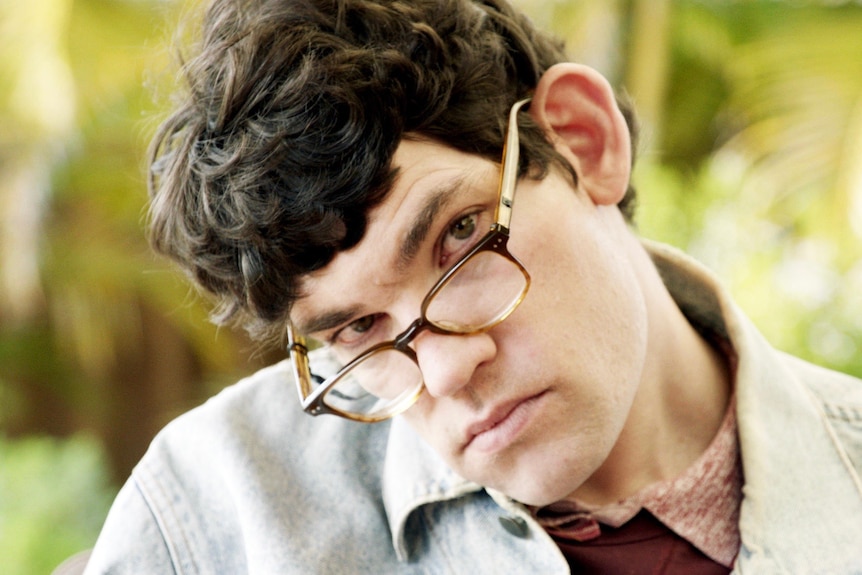 A young man in a collared shirt tilting his head to the side. He wears dark thick-rimmed glasses and looks expressionless 