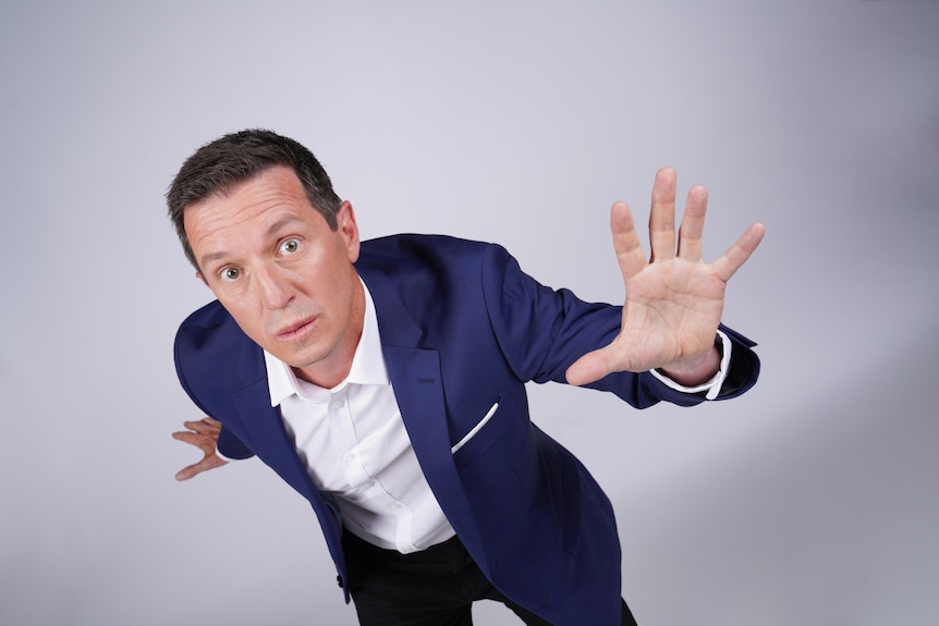 Rove McManus, 52, in a suit, looks bemused, one arm stretching forward, the other reaching back.