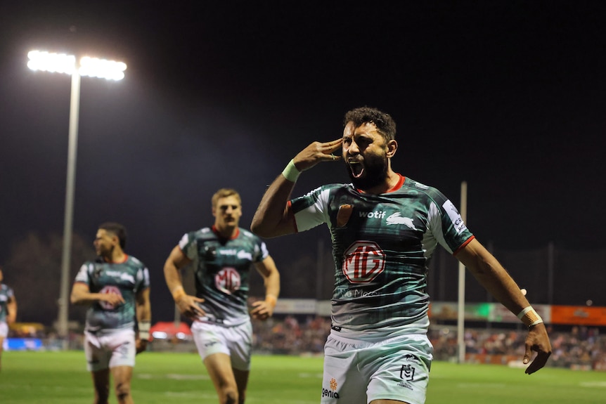 A rugby player screams in celebration and holds his hand to his head as he walks under lights on the pitch with players behind.