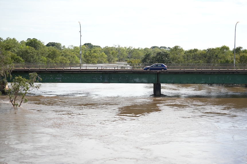A small blue car going across bridge, with river levels very high up on the bridge's standing. 