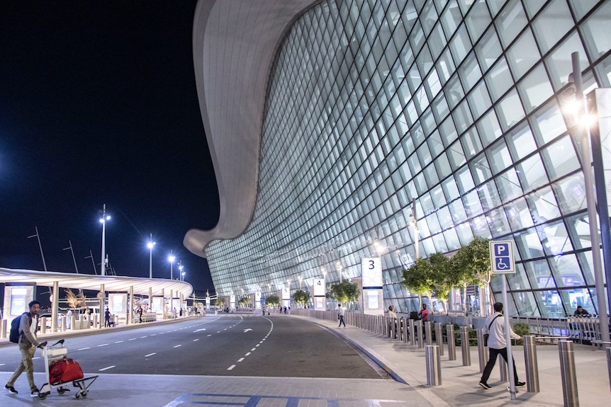 The large glass facade and undulating roof structure of an airport lit up at night.