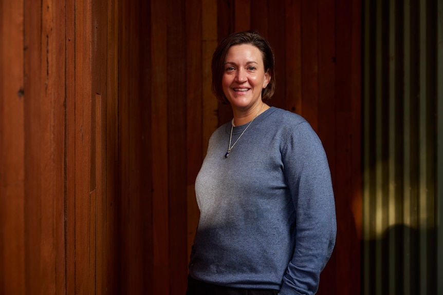 Woman with short brown hair smiles at camera in blue shirt in front of wood wall.