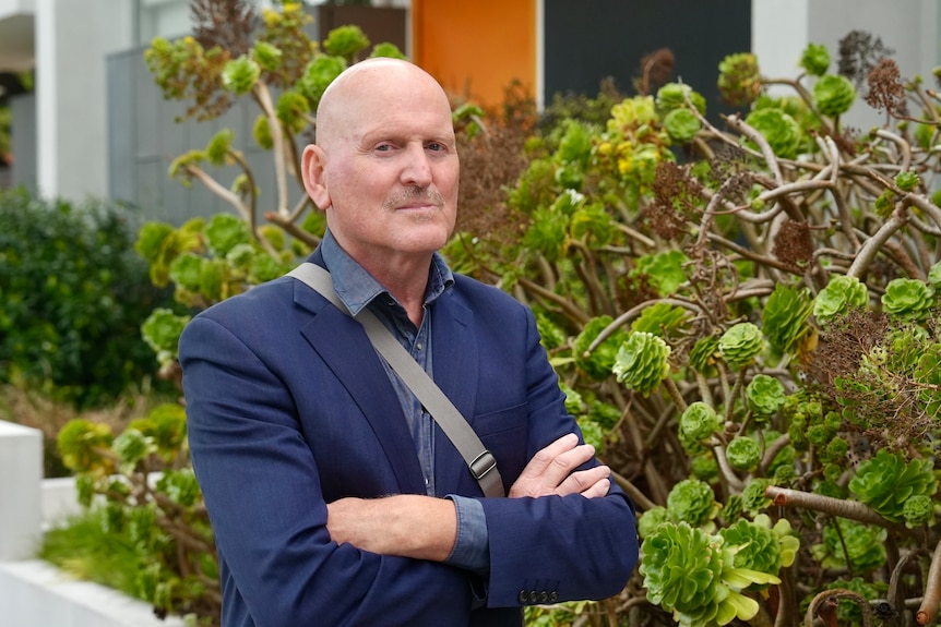 A man in a suit stands outside a garden in front of a house. He has his arms crossed and a serious facial expression.