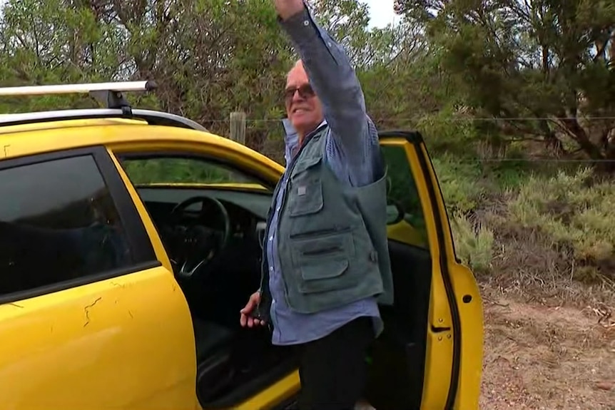 A man smiles and waves goodbye next to his car with the door opened