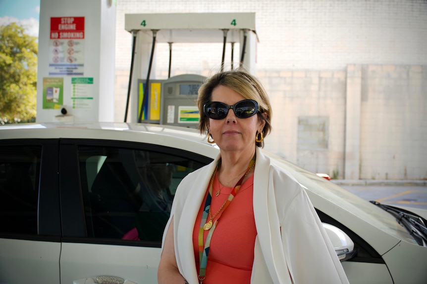 A woman stands in front of her car at a petrol station.