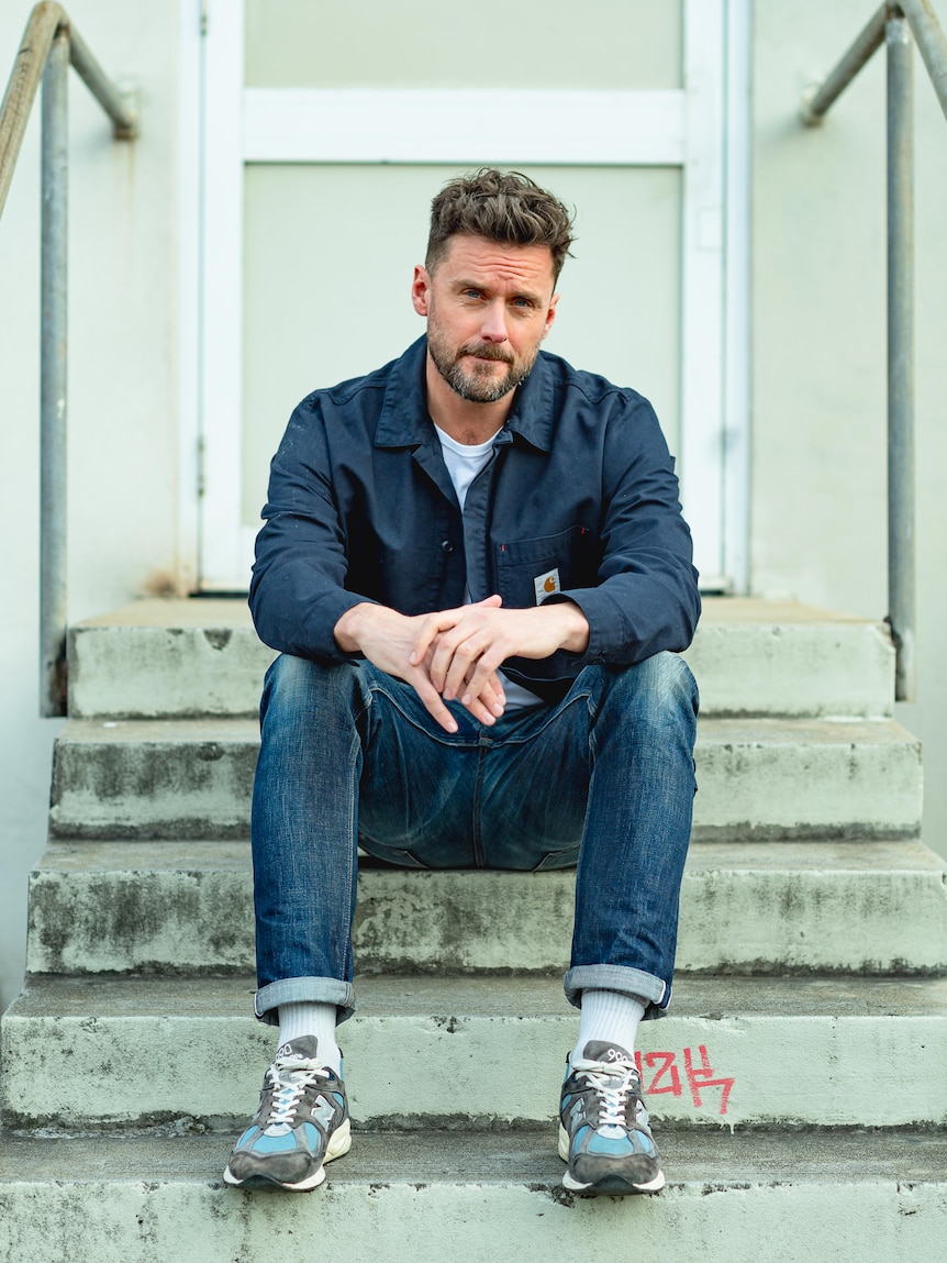 Dave Thornton, 46, bearded, hands clasped, in a jacket and jeans, sits on a stoop with a serious expression.