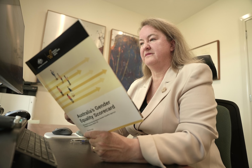 A woman wearing a beige blazer sits at a desk reading a report.