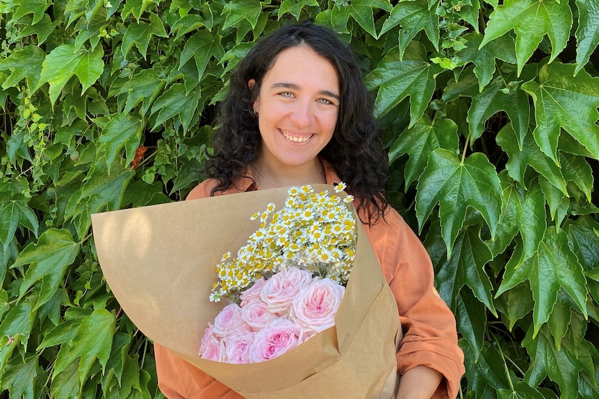 A woman smiling holding a bouquet of pink roses and small white and yellow flowers.