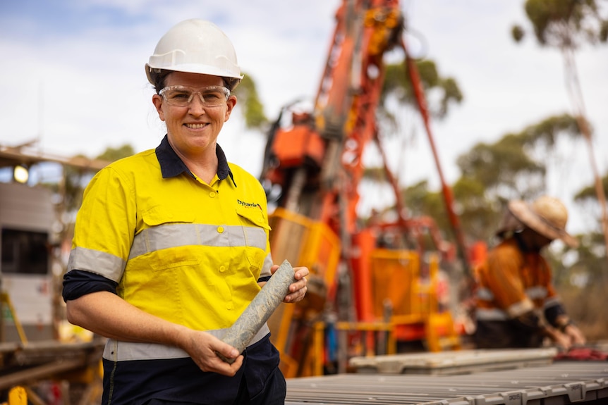 A female geologist holding a drill core specimen next to a drill rig.