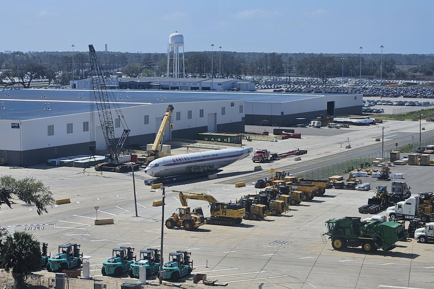 The Boeing 707 sits on a tarmac at an airport surrounded by large machinery and buildings.