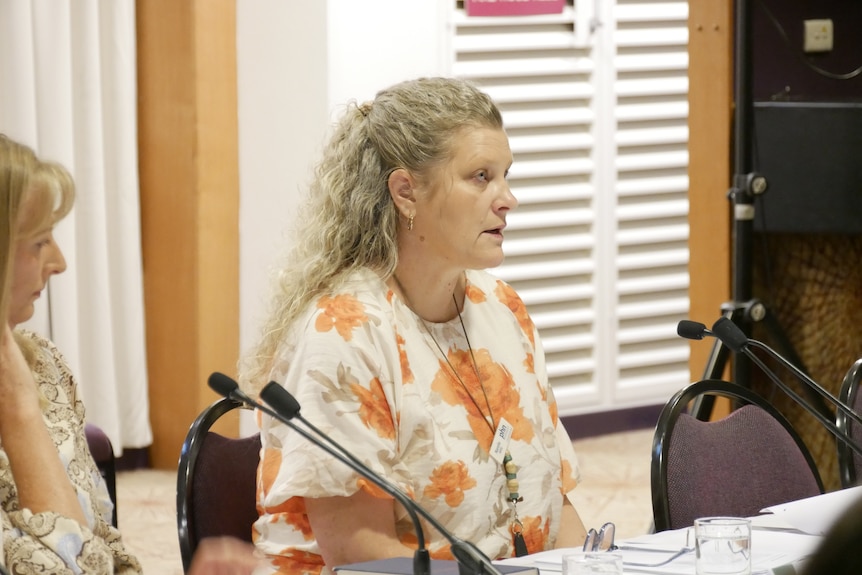 A woman with curly blonde hair sits at a table behind a microphone.
