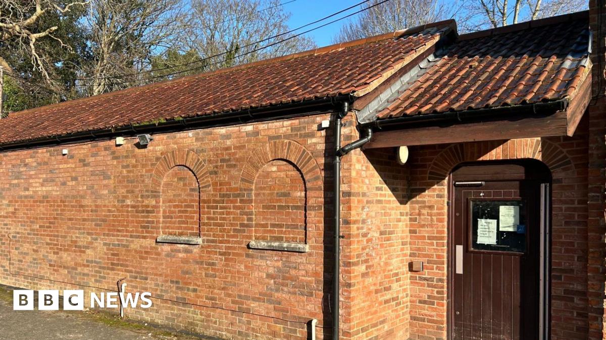A long brick building, with a wooden door at the end. Behind there are trees peeking out above it.