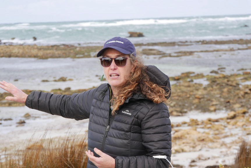 A woman wearing a jacket and glasses speaking on a beach. 