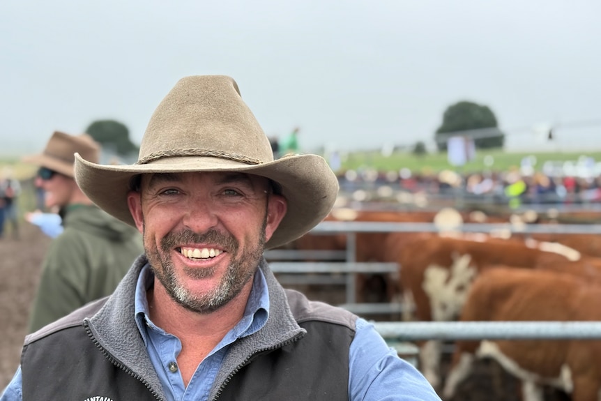 A man wearing an akubra has a big smile on his face, standing in front of a cattle pen