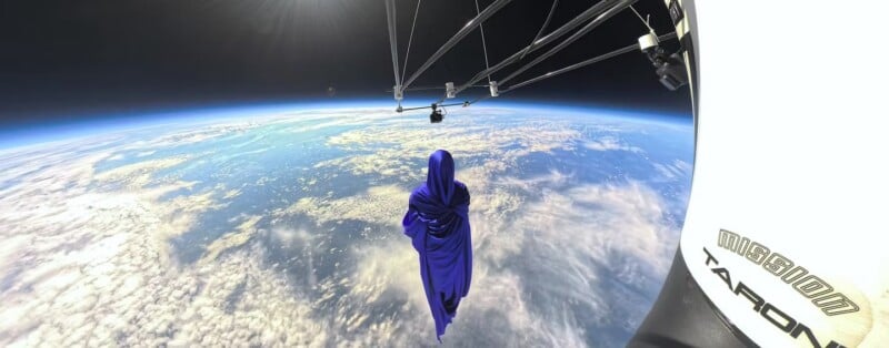 A figure in a blue cloak floats above Earth, tethered to a capsule by cables, with clouds and the planet’s curve visible below against the blackness of space.