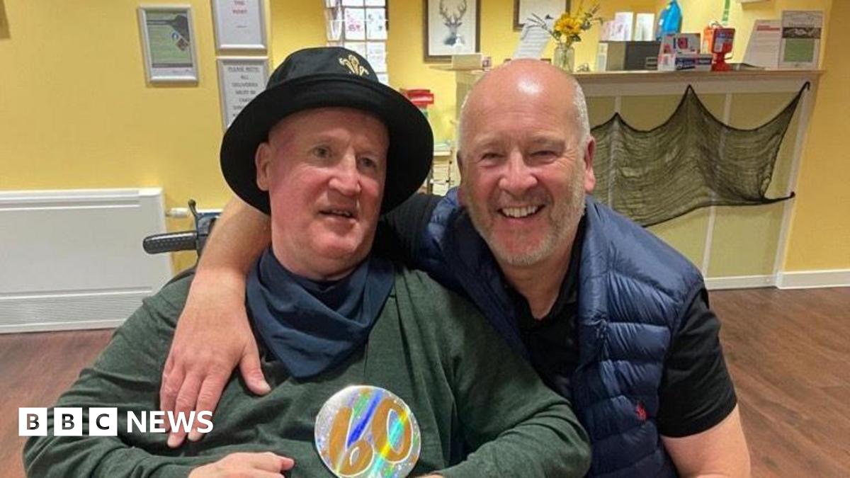 Anthony has grey hair and beard in a blue gilet with a black t-shirt underneath. He has his arm round his brother Keith who is in a wheelchair and is wearing a green long-sleeved t-shirt with a navy bandana round his neck and a black hat on.