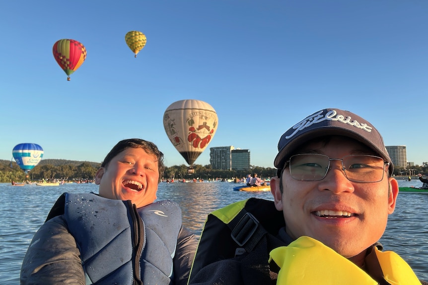 Two men on a kayak on a lake with hot air balloons in the background. 