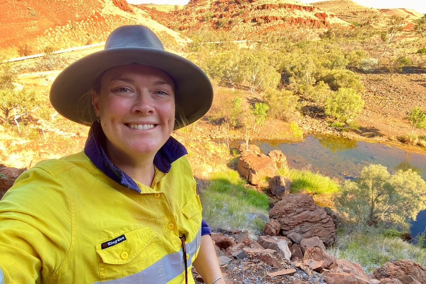 A selfie of a smiling woman in hi-vis against a backdrop of red rocks, spinifex, and a small natural pool of water. 