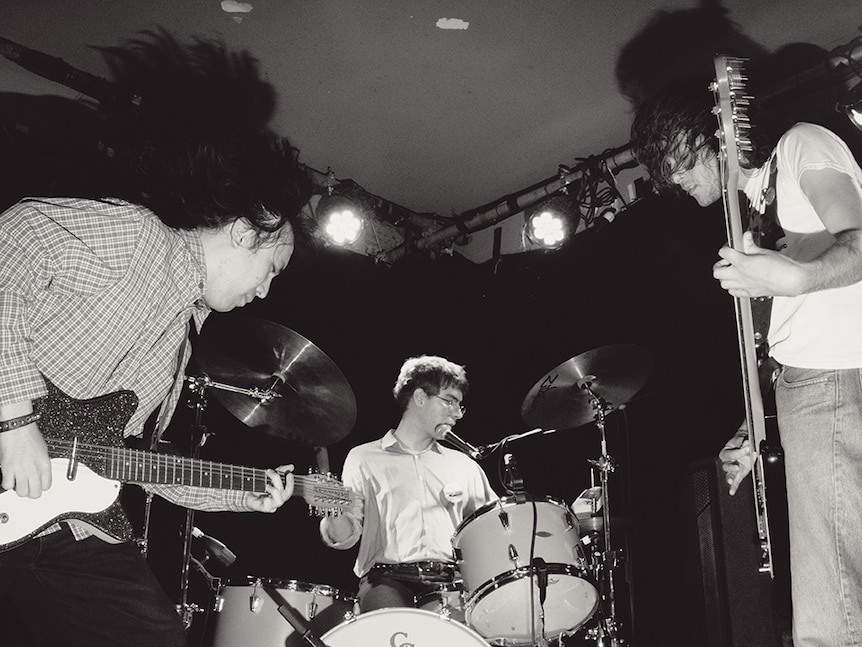 Black and white photo of band on stage, with two guitarists either side of a drummer, all playing their instruments