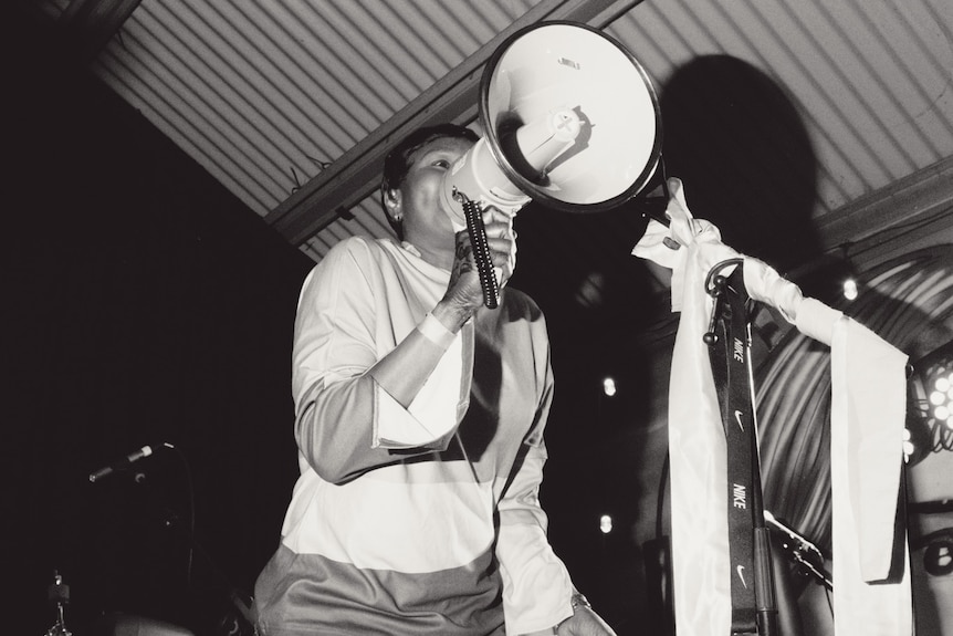 Black and white photo of a woman holding a megaphone on stage behind a microphone