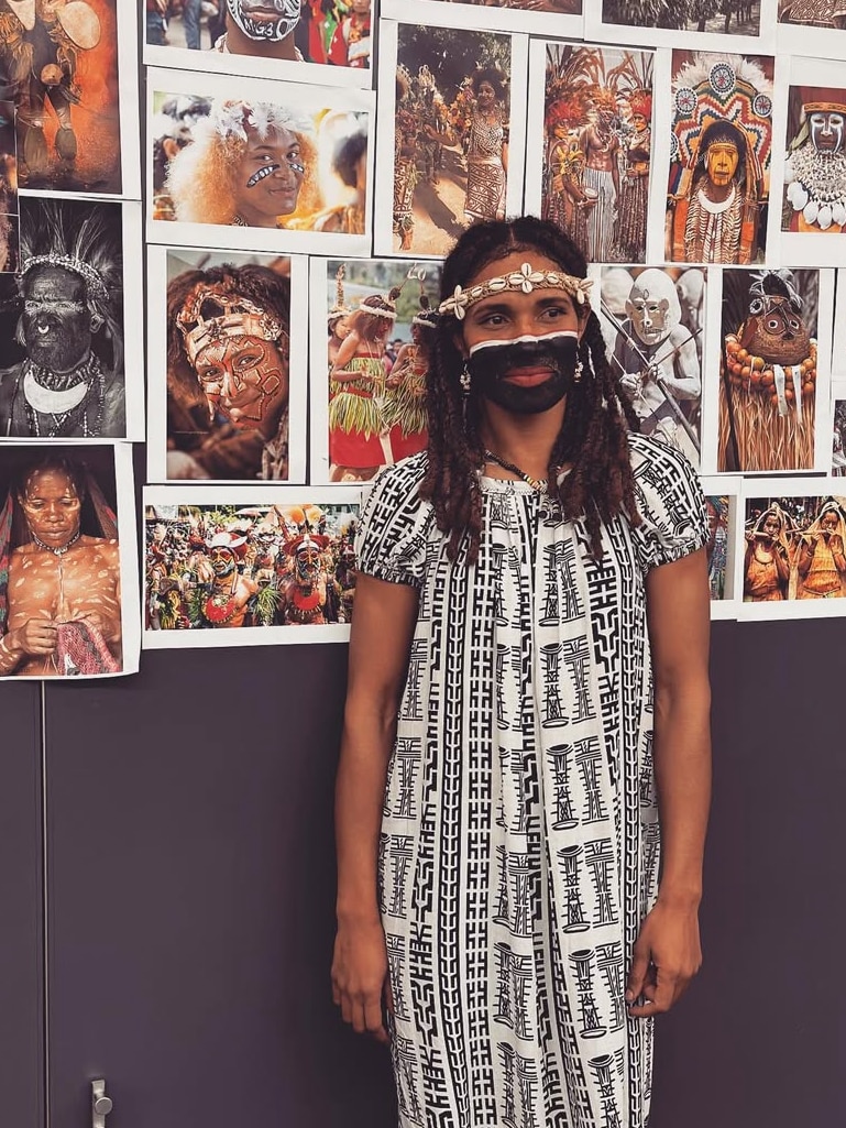 A  young Papua New Guinean woman stands in front of a wall with a number of photos of PNG women.  She wears black face paint.
