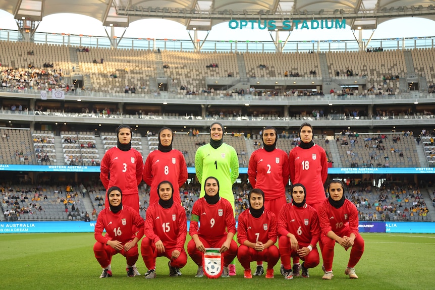 Members of the Iran women's football team are lined up in two rows for a team photo on the pitch.