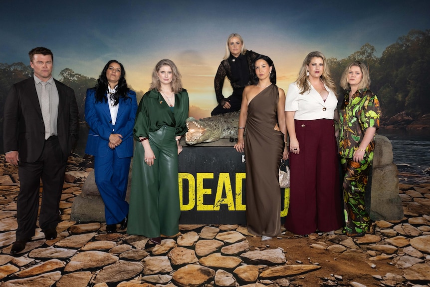 The cast of Deadloch, including Luke Hemsworth and Nina Oyama, pose with a model of a crocodile near a media wall.