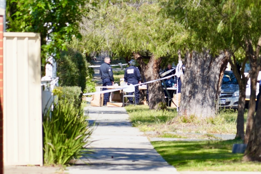 Police forensic officers stand on a footpath outside a house cordoned off by police tape in a suburban street.