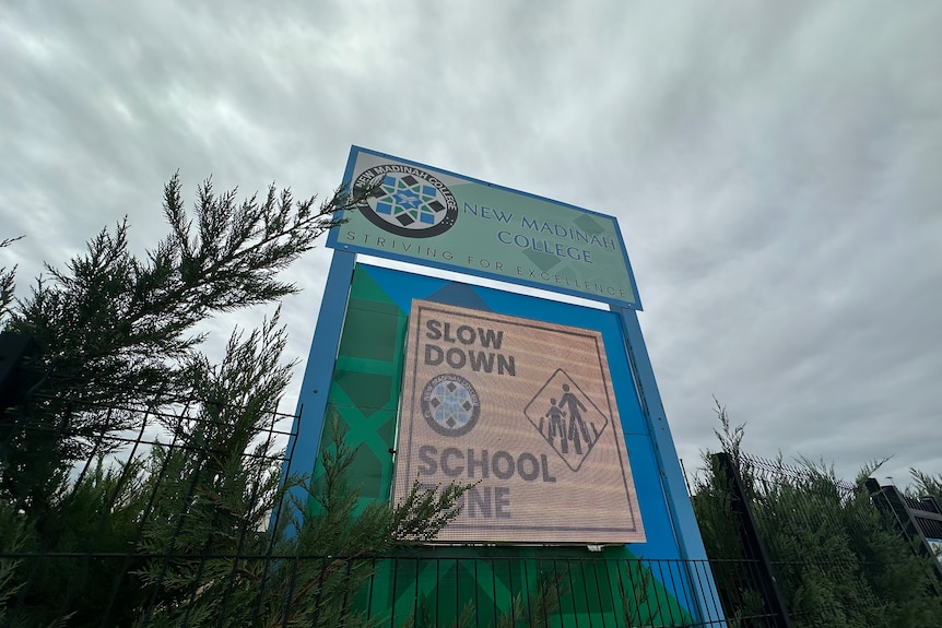A school building with a sign at the entry in front of a carpark. 