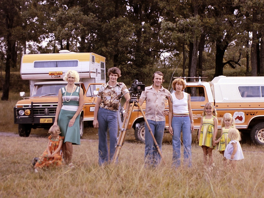 Two men, two women and four children stand in a grass field in a line in 1970s attire. Behind two campervans and a camera centre