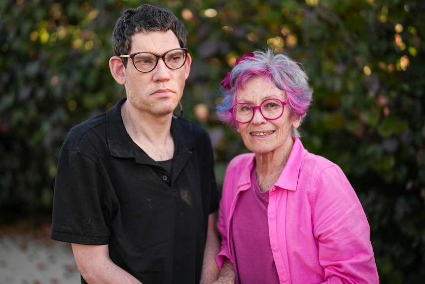 Man aged 45 wears dark-rimmed glasses and black shirt, standing next to his mother who wears all pink outfit, glasses and hair
