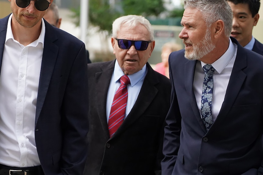 Barry Cable walks between two men in the Perth CBD, wearing a dark suit, red tie and sunglasses.