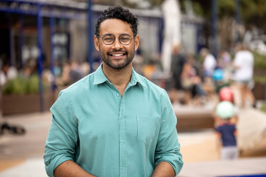 A man with short hair and glasses wearing a green and white striped shirt smiles at the camera outside.