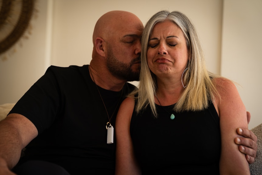 A man and a woman sit on a counch he has a beard and bald head and is kissing her cheek, she is crying.