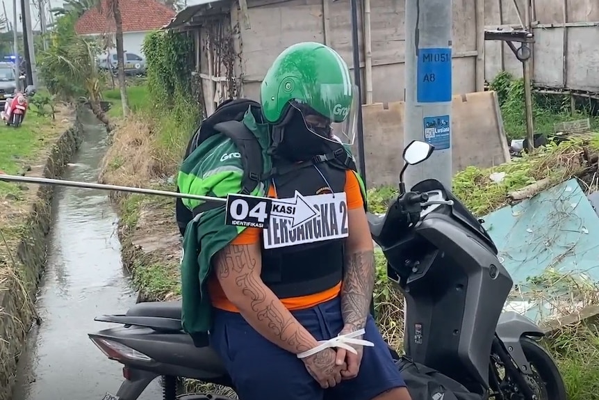 A man with his wrists tied and face covered, wearing a ride-share driver uniform, on a motorbike surrounded by police