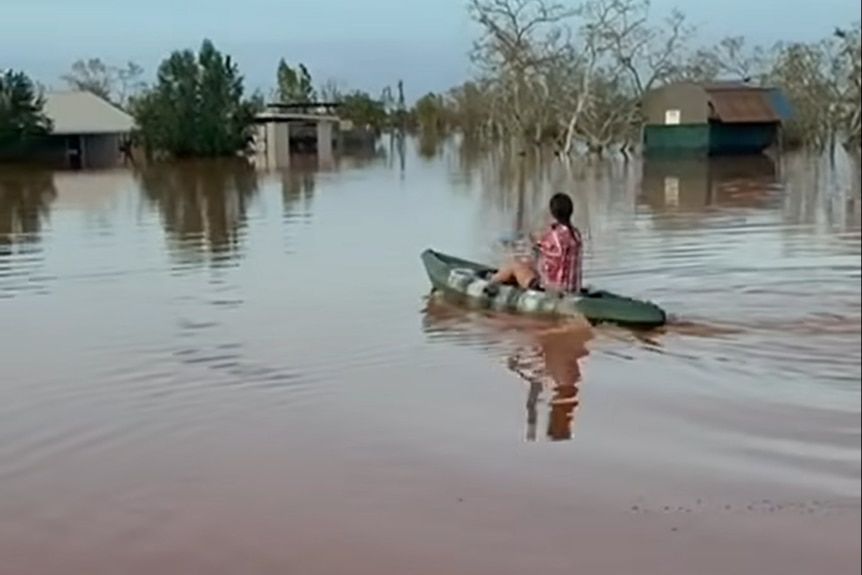 A woman paddle boats through a rural property, with water almost reaching the roof tops of buildings.