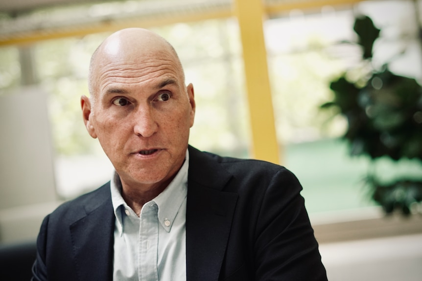 Man in collard shirt and black jacket looks past the camera, with a serious look on his face