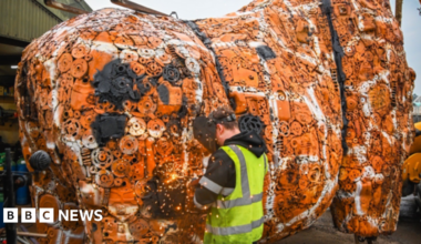 A man with a yellow reflective jacket and face mask facing a large orange and white sculpture, representing the main body of the giraffe, which is made from thousands of car parts.
