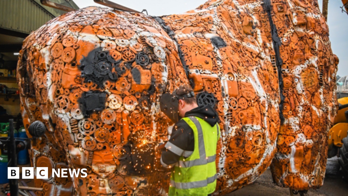 A man with a yellow reflective jacket and face mask facing a large orange and white sculpture, representing the main body of the giraffe, which is made from thousands of car parts.