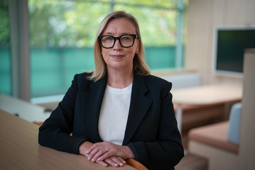 A middle aged white woman in a navy suit jacket. She has glasses and blonde hair