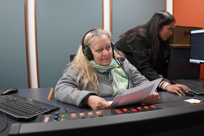 A woman with a microphone headseat sits in a radio studio, holding a piece of paper. 