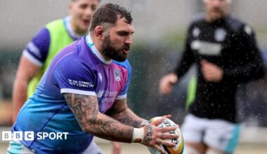 Jamie Bhatti in the purple shirt of Glasgow Warriors holds the ball during a warm-up, with two team-mates indistinct in the background.