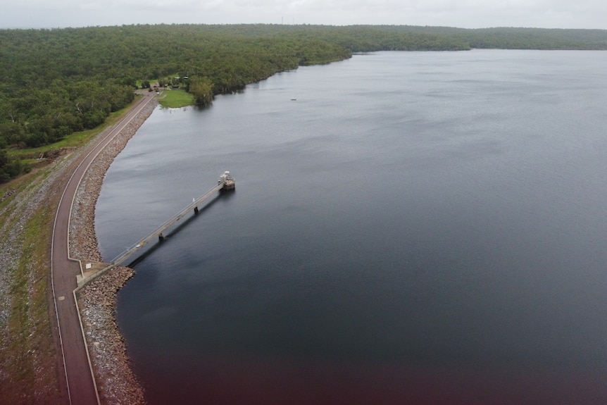 Footage of a full dam with a road on the left and trees in the background