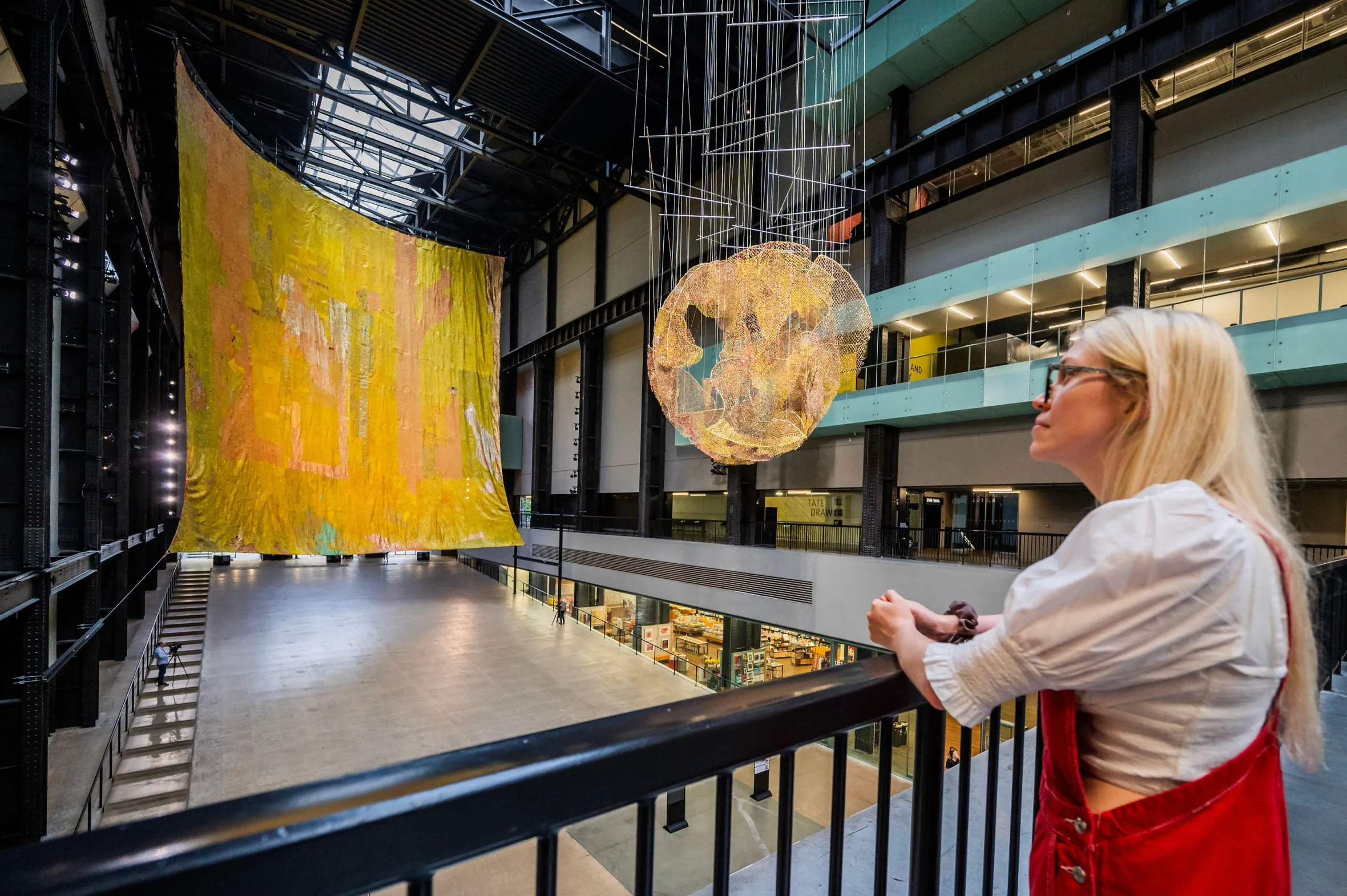 A large yellow and orange fabric artwork hangs from the ceiling in the Tate Modern's Turbine Hall, with a smaller, orb-like metallic sculpture in front of it and a person looking at it.