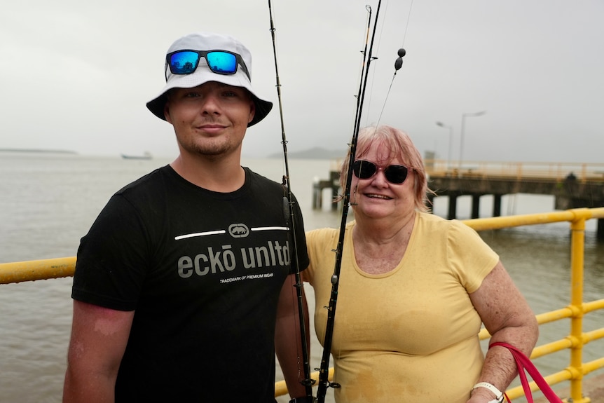 A man and woman standing beside eachother on a jetty holding fishing rods. 