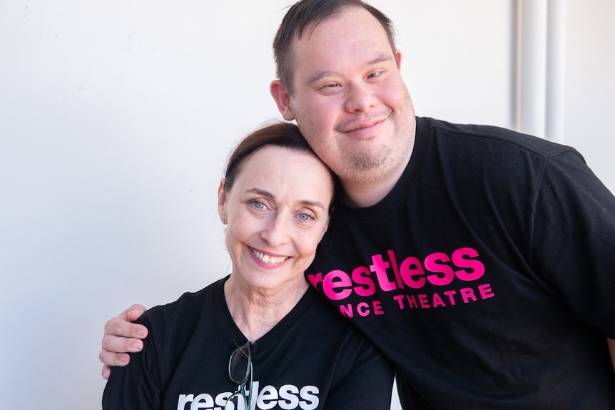 Woman and man side by side, woman is sitting, man has arm around her shoulders, smiling at camera