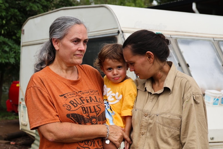 A woman in an orange shirt with grey hair stands beside a woman in a khaki shirt, with a small child held between them