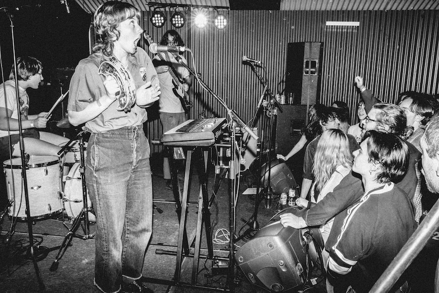 Black and white photo of band on stage, with a female on tambourine next to a drummer and guitarist before a lively stage