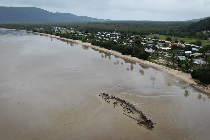 An aerial view with a grey coast to the left, a small series of houses and green mountains in the background.