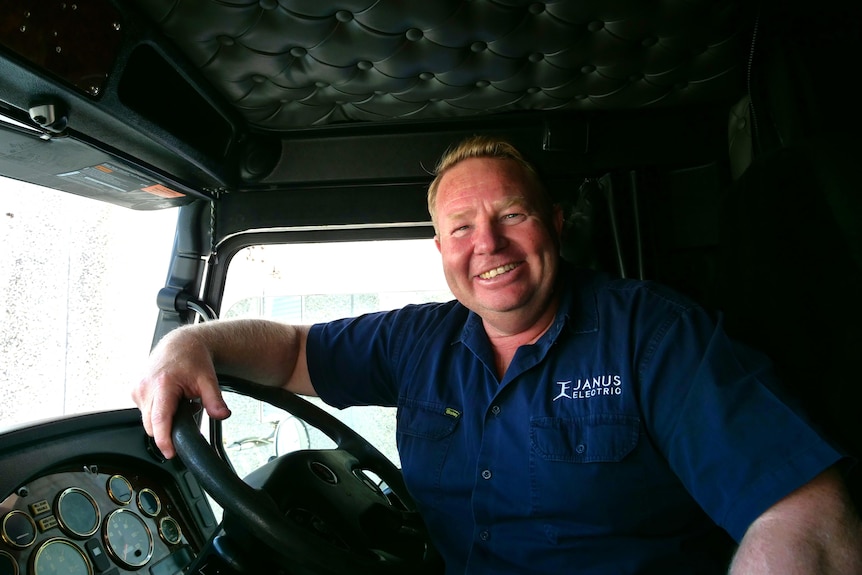 A man sitting behind a steering wheel in a truck cabin. He wears a dark blue shirt and is smiling.